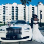 Bride and groom embrace with the bride's flowers resting on the hood of the Rolls Royce Ghost.
