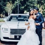 Bride and groom embrace and sip champagne by the Rolls Royce Ghost limousine.