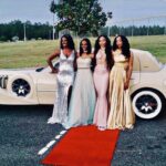 Four women pose in formal dresses in front of Exotic Limo's Excalibur Godfather limousine.