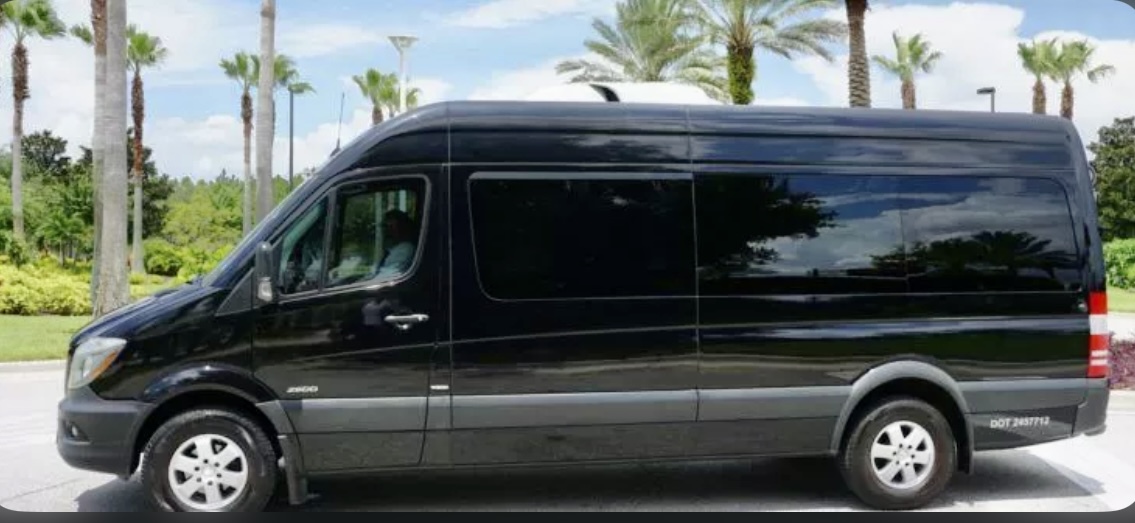 The full side view of our black Mercedes passenger van, spacious enough to accommodate 14 passengers, parked outdoors beside Florida palm trees. The Mercedes van belongs to the Exotic Limo Orlando vehicle fleet. 